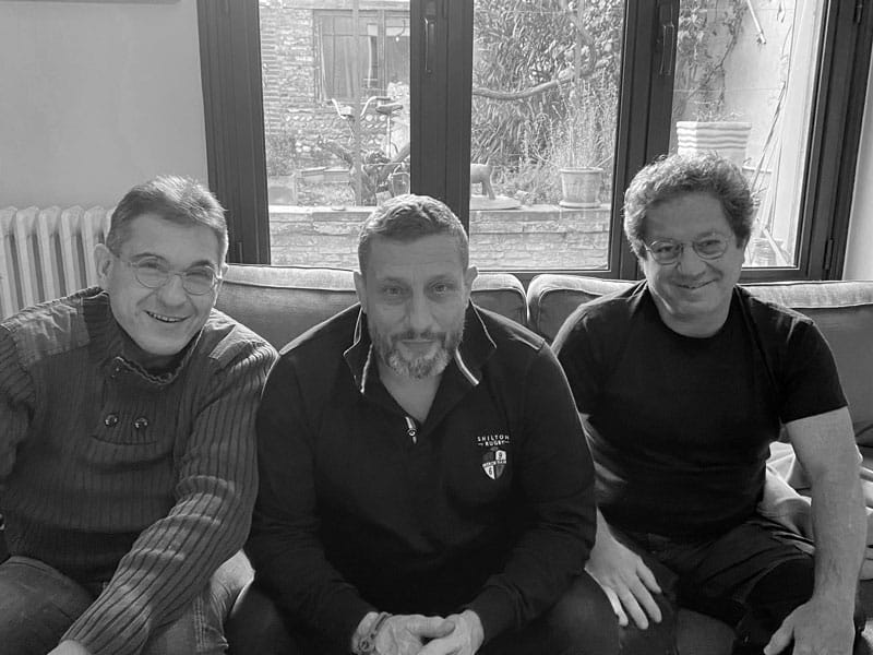 Trois hommes souriants entre amis sur canapé Trois hommes souriants, vêtus décontractée et portant des lunettes, sont assis sur un canapé devant une grande baie vitrée donnant sur un extérieur végétalisé. L'homme central porte un pull noir avec inscription "SHILTON RUGBY".